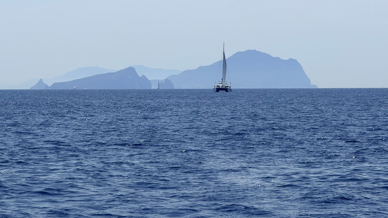 Open sea off the Aeolian islands, a catamaran under sail against layered island silhouettes on the horizon